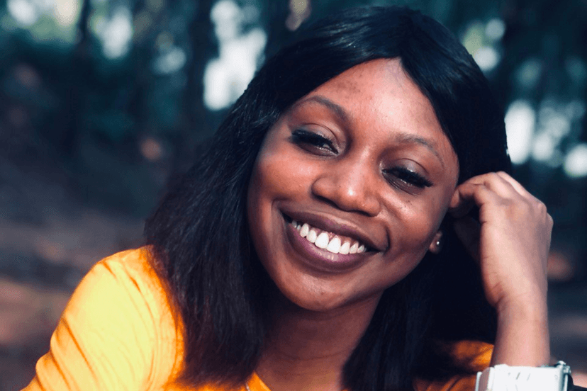 Fortune Aganbi smiling outdoors in a yellow top, one hand resting against her head.